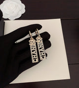 Gold earrings with 'CHANEL' branding held by a black-gloved hand on a white surface.