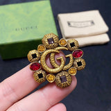 Gold brooch with red and yellow gemstones held by a hand, Gucci box in the background